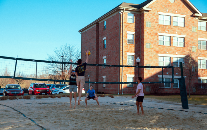 People play volleyball on a sand court. There is a building in the background.