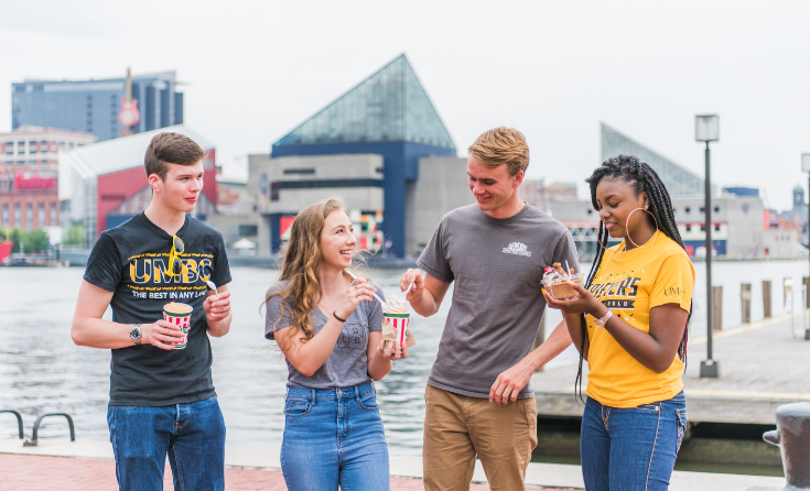 Four people standing in the Baltimore Inner Harbor and eating Rita's Italian ice.
