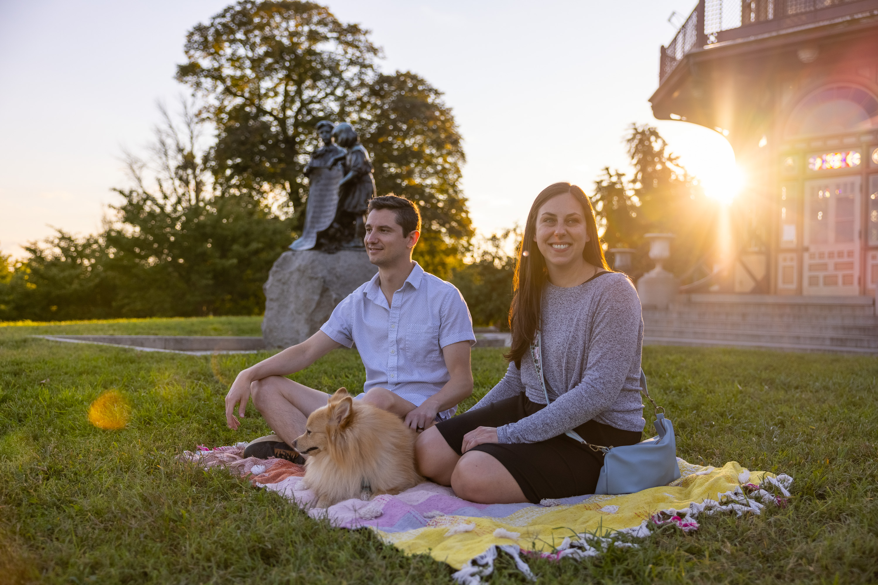 Photograph shows a man and a woman sitting on a blanket in the grass in a park with a dog.