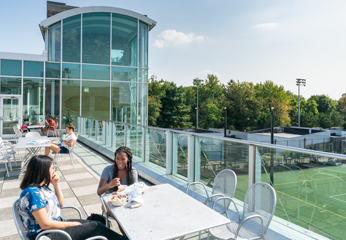 People sit at tables on rooftop of a building overlooking an athletics field.