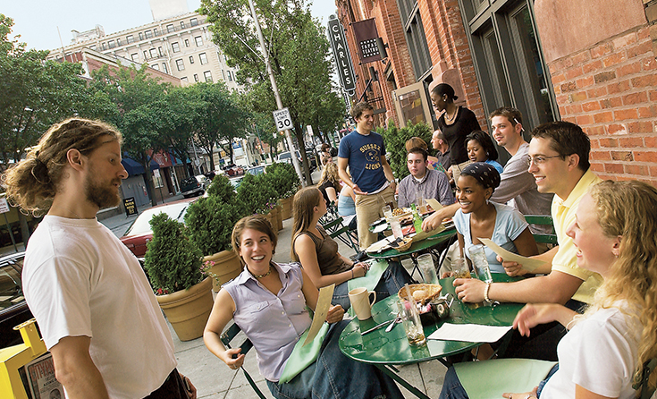 People sit at a table outside at a restaurant while another person, standing, talks to them.