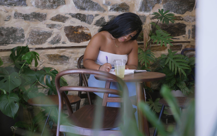 Person sits at table and writes in notebook. They are in front of a stone wall and surrounded by plants.