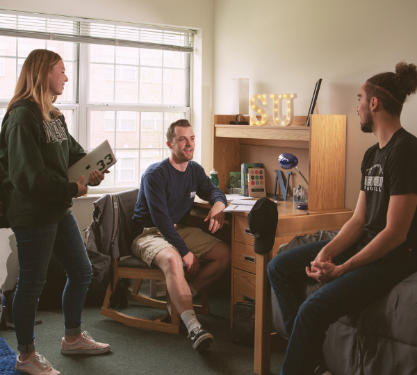 Two people sit, and one person stands in a dorm room talking.