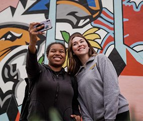 Two people pose for a selfie in front of a mural.