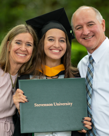 A person in graduation attire holds a diploma cover that says Stevenson University and poses with two other people.
