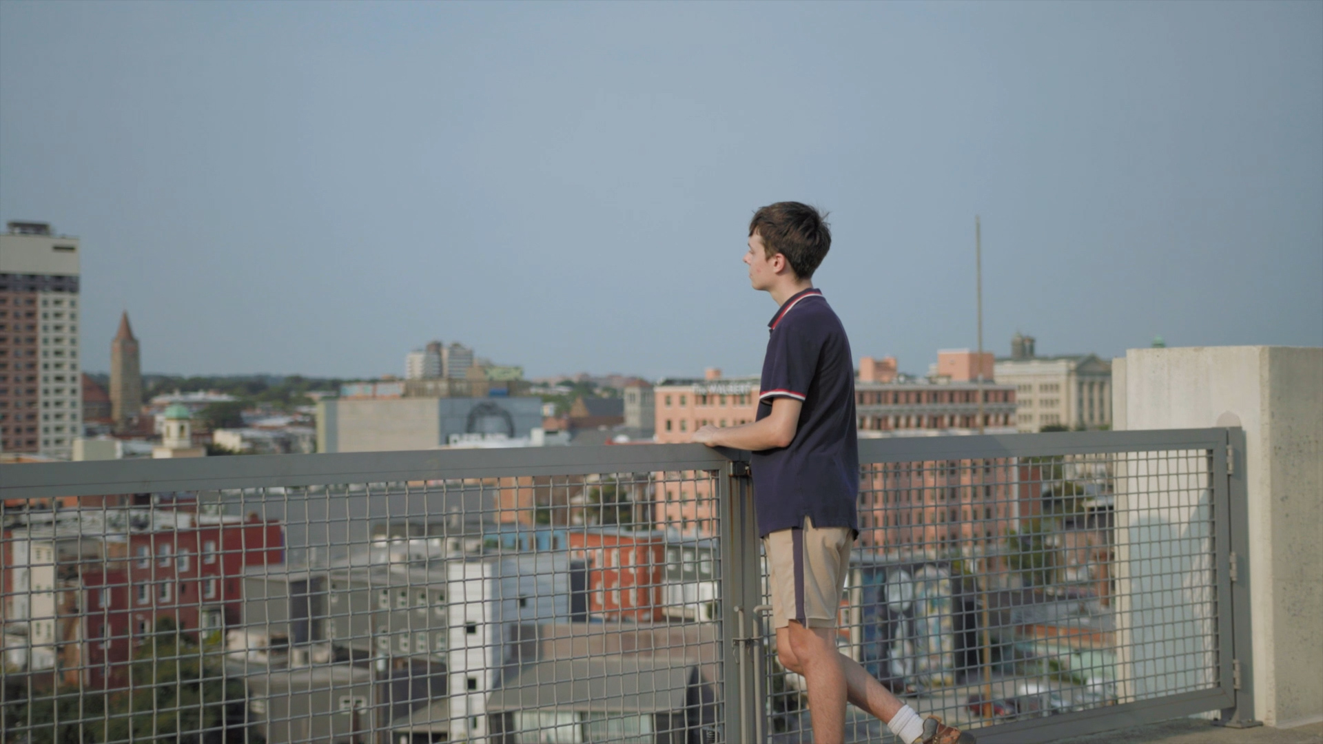 Person stands against wire fence on rooftop and looks out over a city.