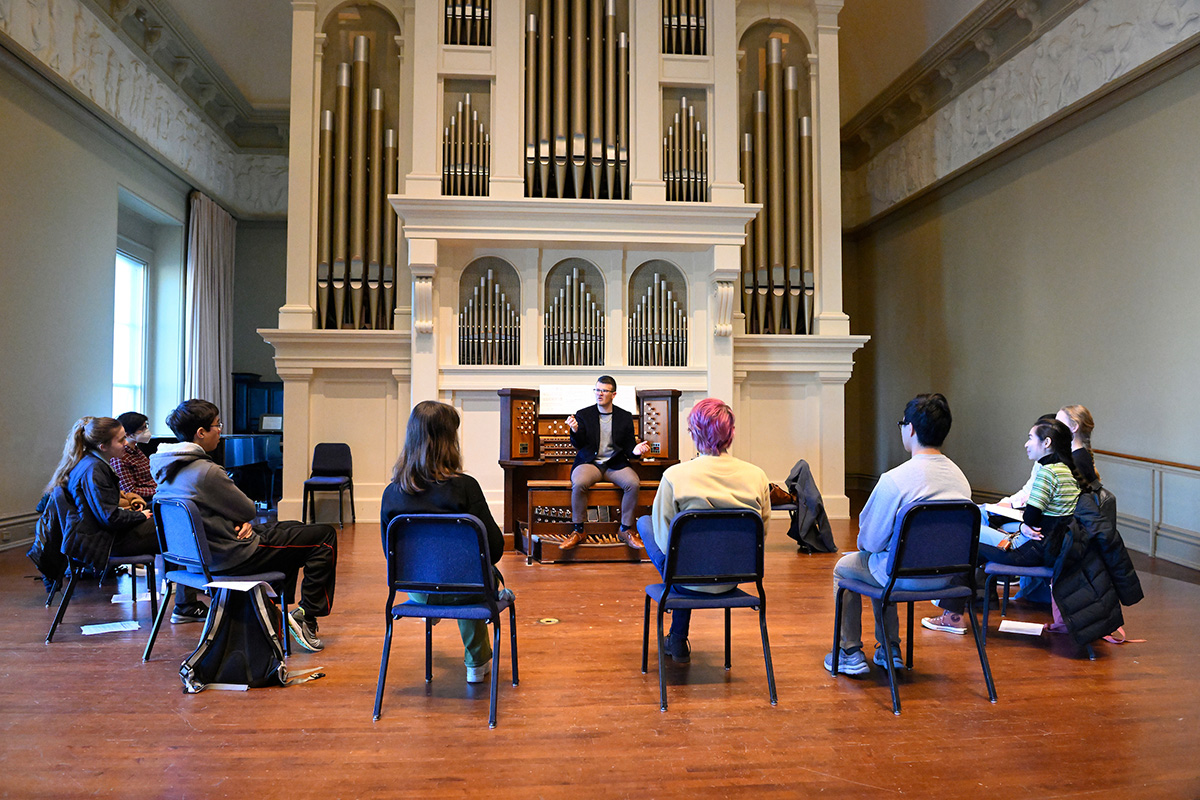 Hopkins students listening to a lecture on a stage in front of an organ.